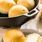 A plate with a halved bread roll, one half topped with a slice of butter, sits in front of a cast iron pan filled with whole no knead rolls.