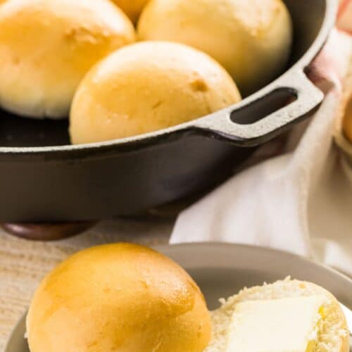 A plate with a halved bread roll—one half topped with a pat of butter—sits in front of a cast iron skillet filled with golden No Knead Rolls.