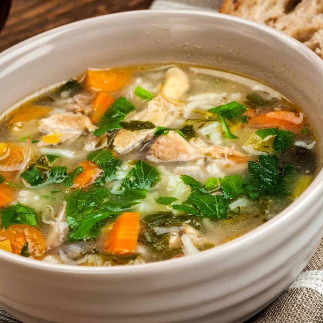 A bowl of Turkey Rice Soup with pieces of turkey, carrots, celery, rice, and fresh herbs, served with a slice of bread in the background.