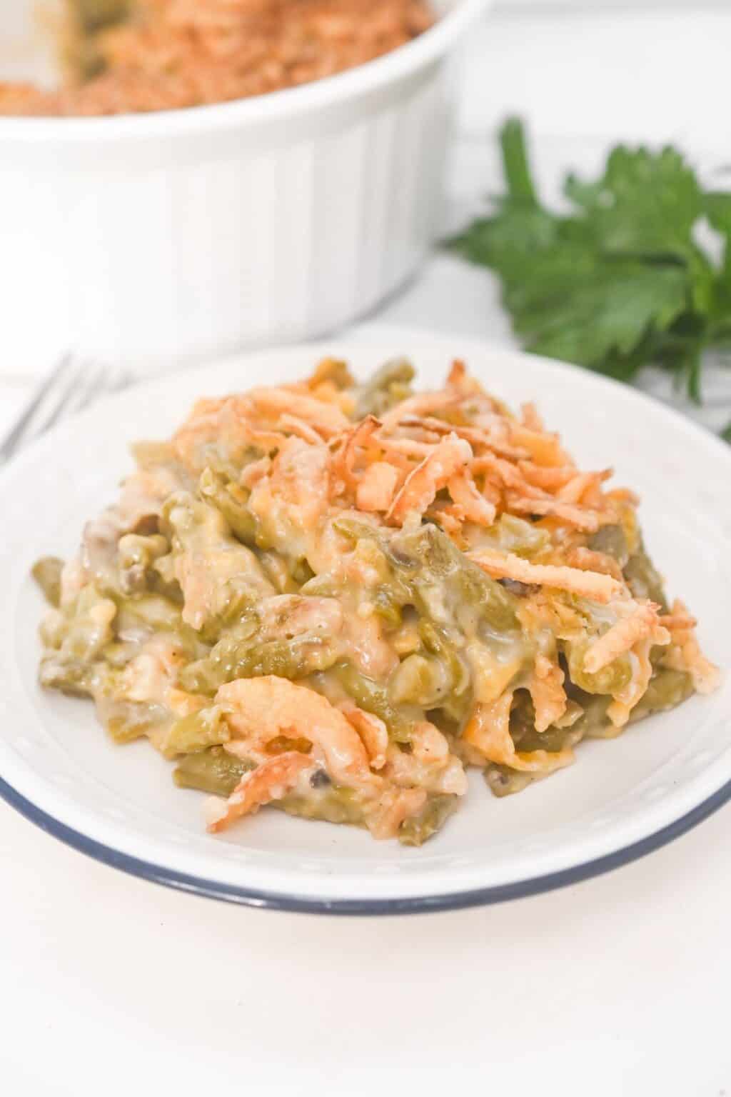 A serving of French’s Green Bean Casserole with crispy fried onions sits on a white plate, with the casserole dish and fresh parsley in the background.
