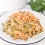 A serving of French’s Green Bean Casserole with crispy fried onions sits on a white plate, with the casserole dish and fresh parsley in the background.