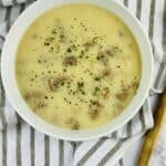 A white bowl of creamy sauce garnished with herbs, placed on a striped towel with a wooden-handled spoon beside it.