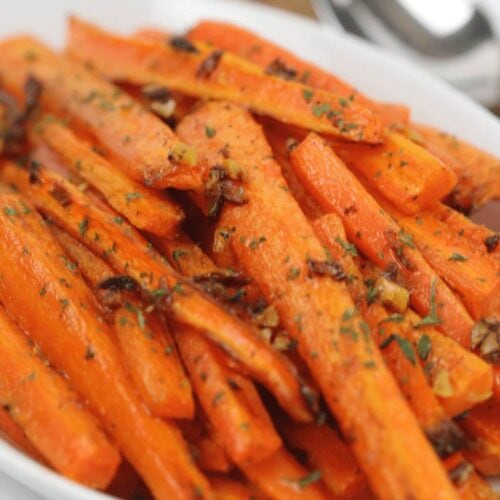 A white dish filled with roasted baby carrots garnished with chopped herbs and bits of caramelized garlic.