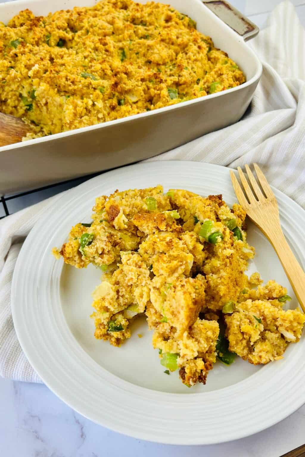 A plate with a serving of Jiffy Cornbread Dressing sits beside a baking dish filled with the same flavorful stuffing, with two wooden forks placed on the plate.