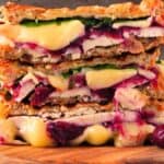 A close-up of a stacked Turkey Panini with melted cheese, sliced turkey, cranberries, spinach, and grilled bread on a wooden board.