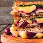 Two halves of a Turkey Panini with melted cheese, spinach, and cranberry sauce are stacked on a wooden board, with grapes nearby.