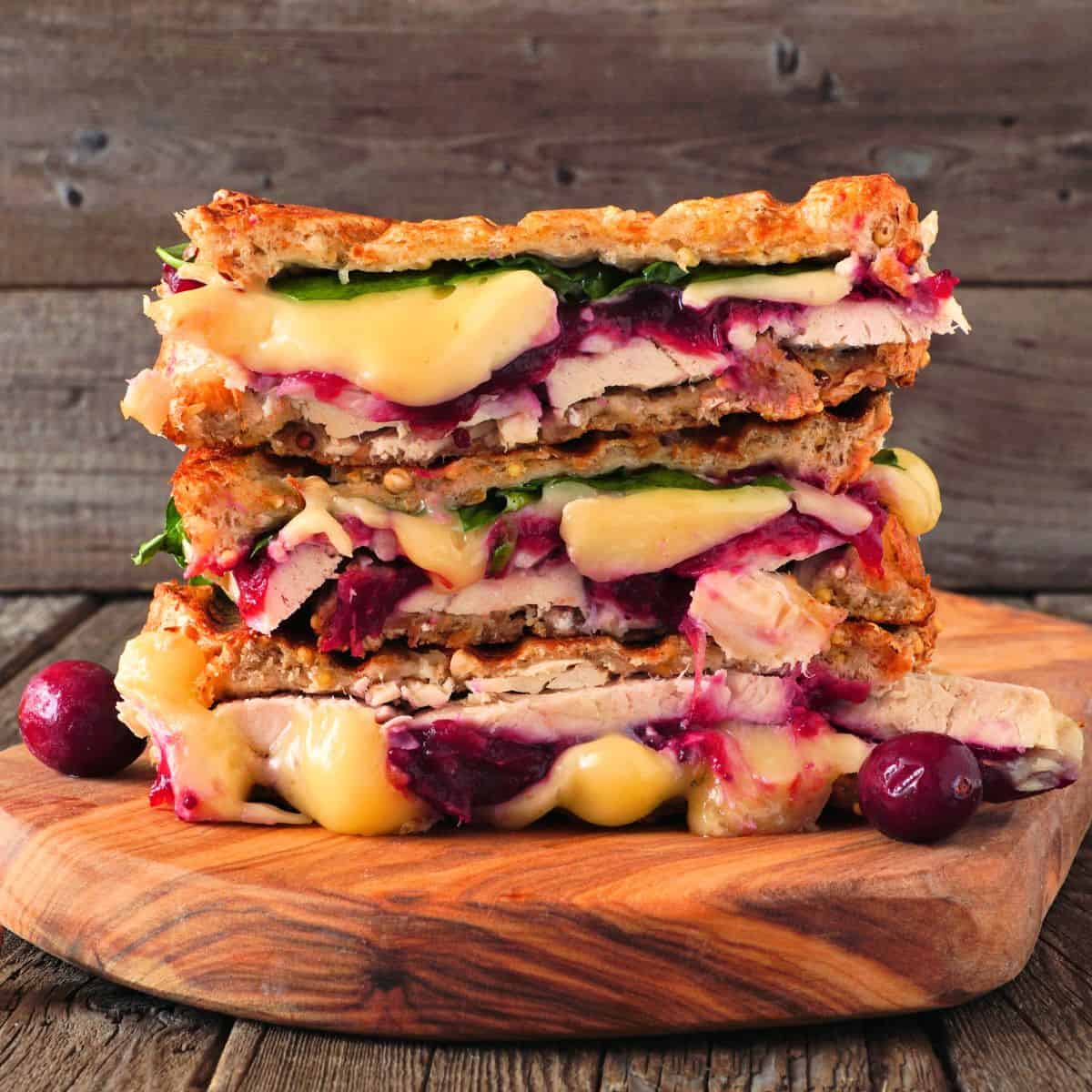 A Turkey Panini stacked with melted cheese, sliced turkey, leafy greens, and cranberry sauce sits on a wooden cutting board, garnished with whole cranberries.