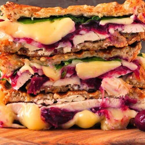 Stacked Turkey Panini sandwiches filled with melted cheese, spinach, and cranberry sauce rest on a wooden board, set against a rustic wooden background.