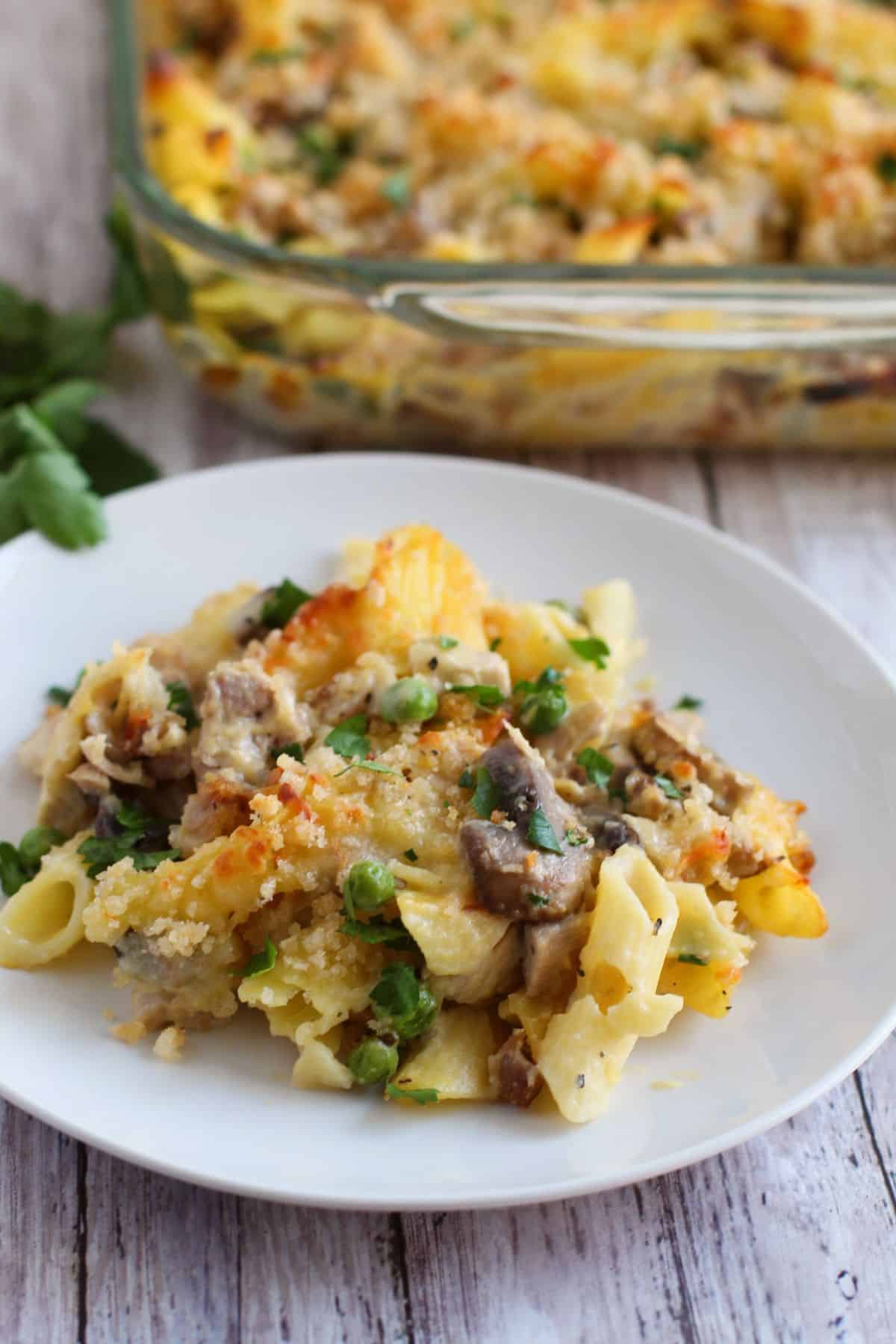A serving of baked turkey pasta casserole with mushrooms and green herbs on a white plate, with a glass baking dish of this delicious Turkey Pasta Recipe in the background.