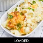 A delectable cheesy cauliflower casserole rests on a wooden surface, its golden melted cheese glistening under fresh green herbs. In the background, another dish and fresh green onions add to the inviting scene.