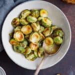 A white bowl filled with Maple Glazed Brussel Sprouts and a silver spoon, on a dark surface next to a gray napkin and a glass of orange liquid.