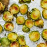 Maple Glazed Brussel Sprouts halves roast on a sheet of aluminum foil, some browned and crisp, with a serving spoon partially visible in the top left corner.
