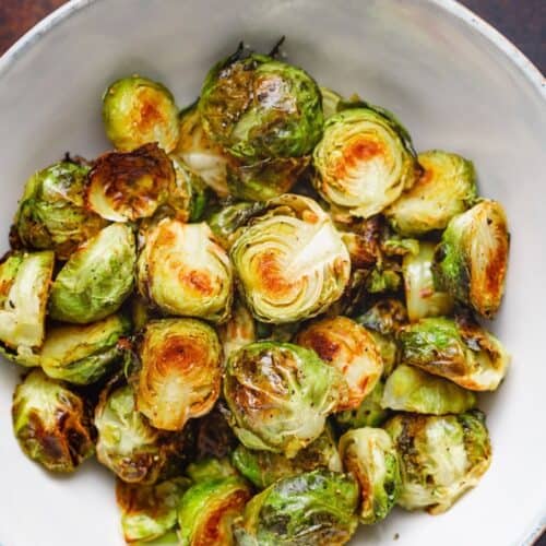 A white bowl filled with Maple Glazed Brussel Sprouts, some halved and browned, set on a dark surface.