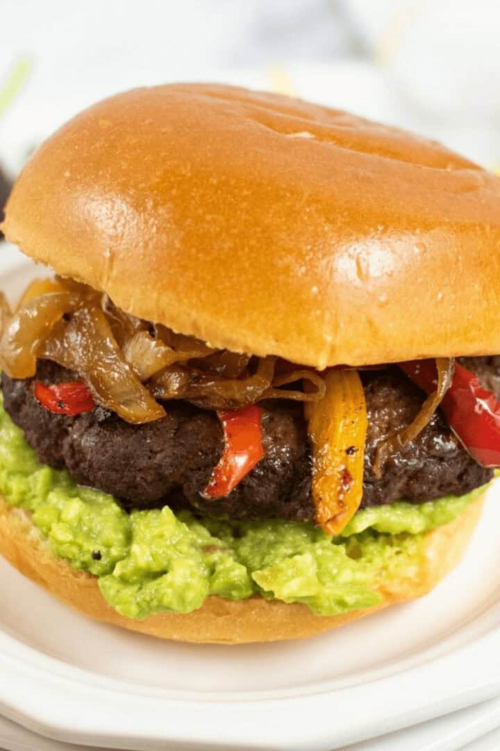 A Mexican Burger with caramelized onions, red and yellow bell peppers, and guacamole on a brioche bun.