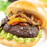 Close-up of the sandwich with a beef patty, guacamole, and sautéed onions and bell peppers on a bun. An avocado is blurred in the background.
