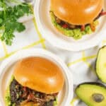 Two gourmet Mexican burgers with caramelized onions and bell peppers on brioche buns, placed on white plates with guacamole spread. Avocados and fresh parsley are arranged beside them.