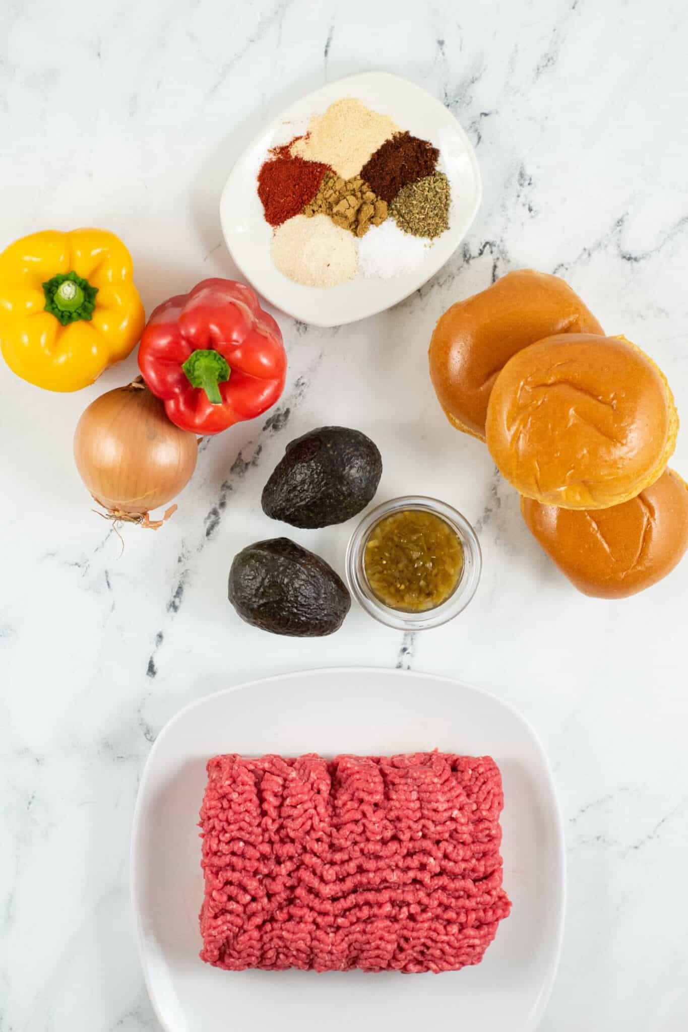 Ingredients for cooking, including ground beef, yellow and red bell peppers, an onion, avocados, buns, a jar of green sauce, and a plate of mixed spices perfect for making a Mexican Burger, all arranged on a marble surface.