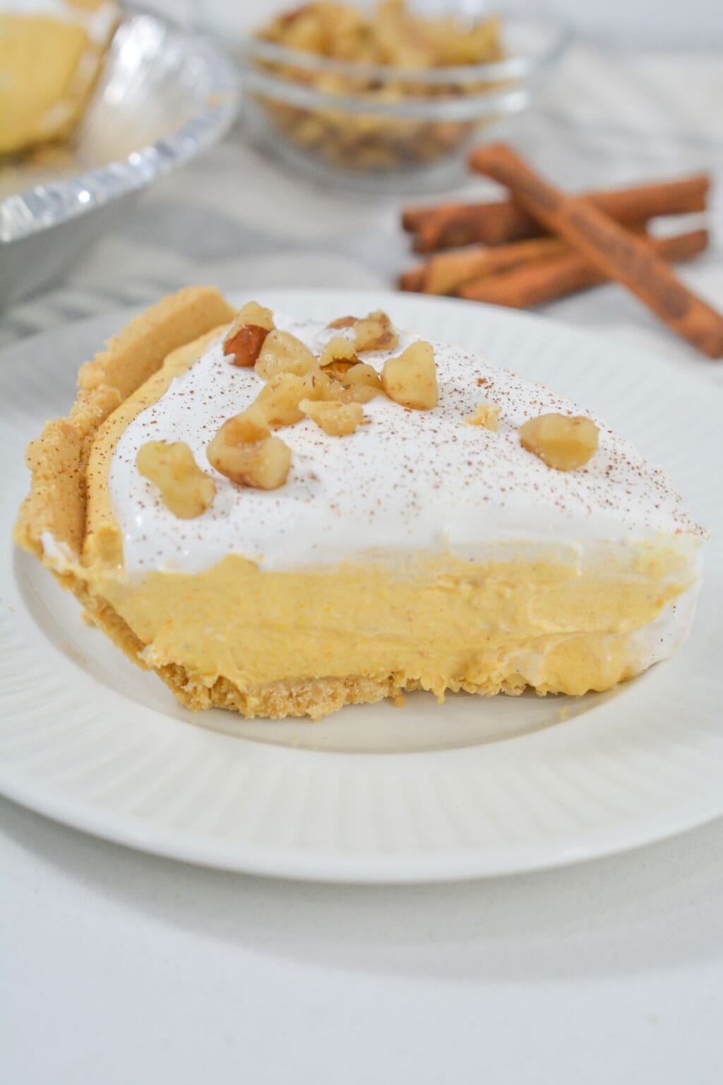 A slice of creamy Pumpkin Cheesecake Pie with a crumb crust, topped with whipped cream and chopped nuts, sits on a white plate; cinnamon sticks and a dish of nuts are in the background.