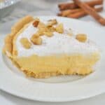 A slice of creamy Pumpkin Cheesecake Pie with a crumb crust, topped with whipped cream and chopped nuts, sits on a white plate; cinnamon sticks and a dish of nuts are in the background.