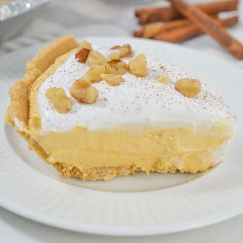 A slice of creamy Pumpkin Cheesecake Pie with a crumb crust, topped with whipped cream and chopped nuts, sits on a white plate; cinnamon sticks and a dish of nuts are in the background.