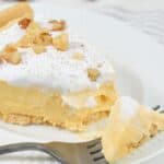 A slice of creamy Pumpkin Cheesecake Pie topped with whipped cream and chopped nuts sits on a white plate, with a fork holding a bite-sized piece. Cinnamon sticks add warmth in the background.