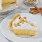 A slice of creamy Pumpkin Cheesecake Pie with whipped topping and chopped nuts on a white plate, with the rest of the pie, a bowl of nuts, and cinnamon sticks in the background.