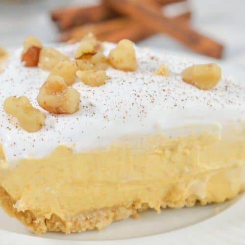 A slice of creamy layered Pumpkin Cheesecake Pie topped with whipped cream, chopped walnuts, and a sprinkle of cinnamon on a white plate.