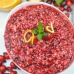 A bowl of Ocean Spray Cranberry Relish Recipe is garnished with mint, lemon peel, and cranberries, with orange halves and fresh cranberries in the background.