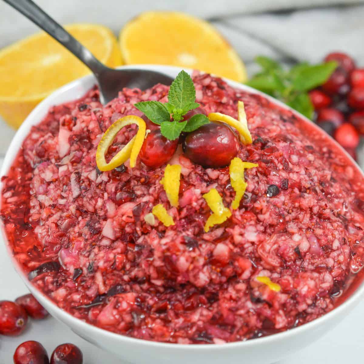 A bowl of finely chopped Ocean Spray Cranberry Relish Recipe garnished with orange peel, cranberries, and mint, with halved oranges and loose cranberries in the background.