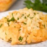 A serving of Old Fashioned Baked Macaroni and Cheese garnished with chopped parsley sits on a white plate, with a casserole dish and fresh parsley in the background.
