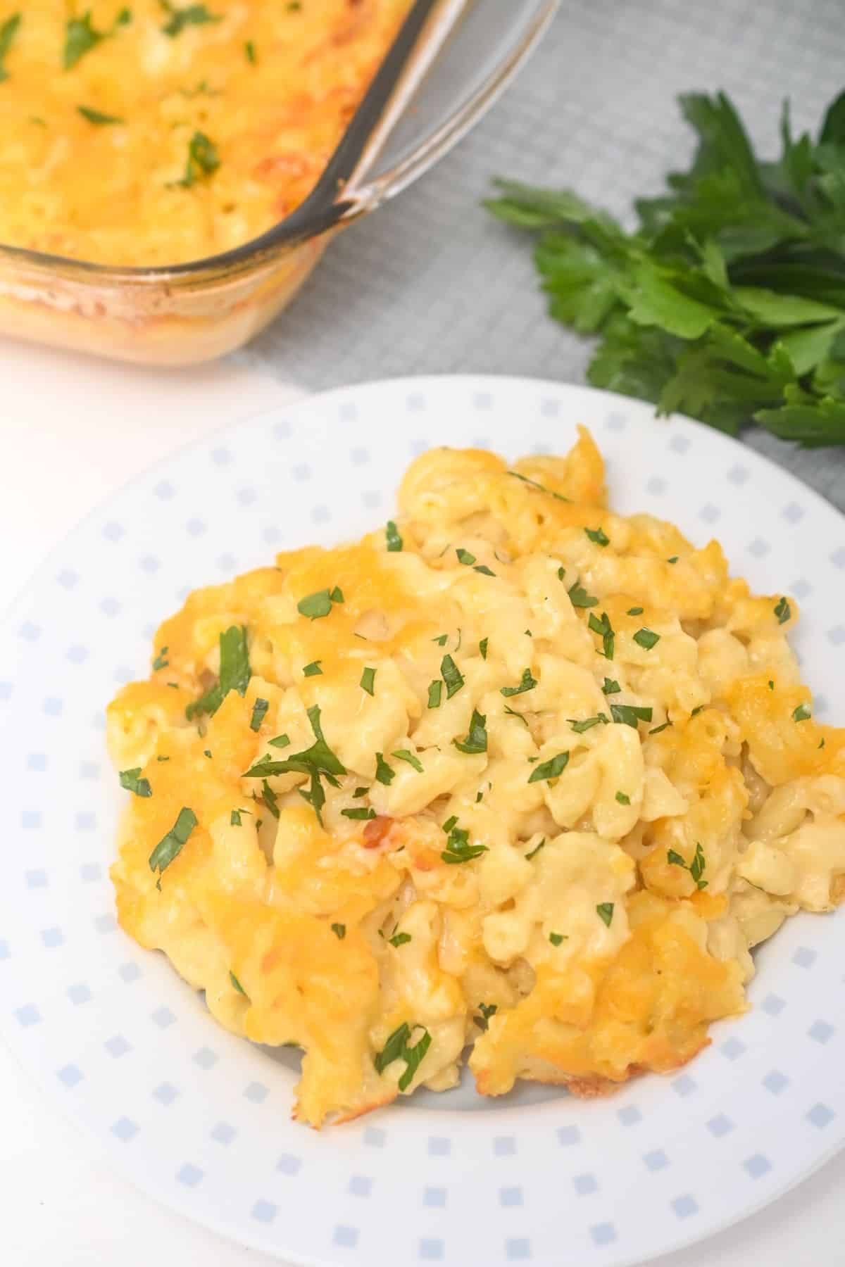 A serving of Old Fashioned Baked Macaroni and Cheese garnished with chopped parsley sits on a white plate, with a baking dish and fresh parsley in the background.