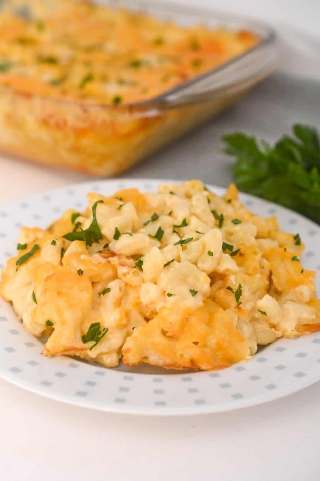 A serving of Old Fashioned Baked Macaroni and Cheese garnished with chopped parsley sits on a white plate, with a casserole dish of mac and cheese in the background.
