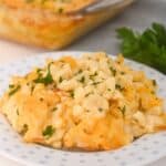 A serving of Old Fashioned Baked Macaroni and Cheese garnished with chopped parsley sits on a white plate, with a casserole dish of mac and cheese in the background.