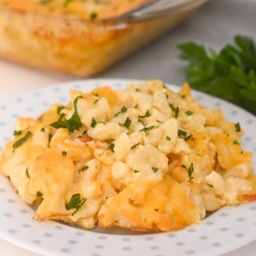 A serving of Old Fashioned Baked Macaroni and Cheese garnished with chopped parsley sits on a white plate, with a casserole dish of mac and cheese in the background.