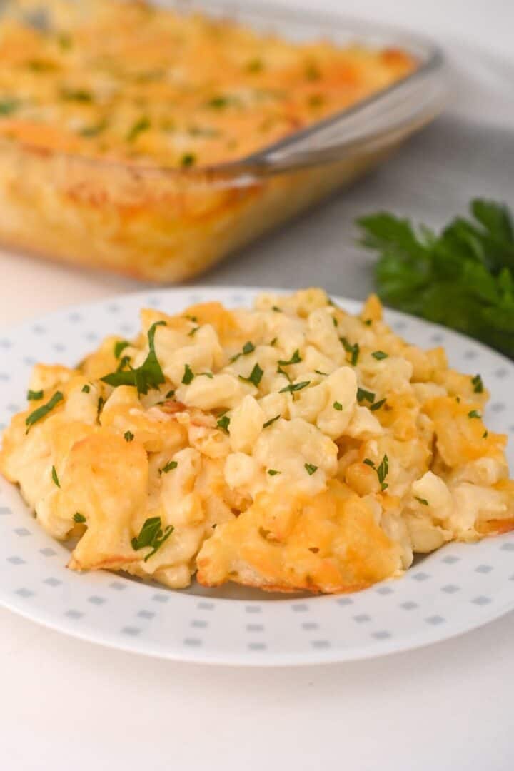 A serving of Old Fashioned Baked Macaroni and Cheese garnished with chopped parsley sits on a white plate, with a casserole dish of mac and cheese in the background.
