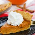 A slice of Old Fashioned Pumpkin Pie topped with whipped cream sits on a black plate, alongside a fork, cinnamon sticks, and a pumpkin in the background.