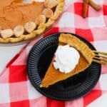 A slice with whipped cream on a black plate, next to a whole pie and two cinnamon sticks atop a red and white checkered cloth.