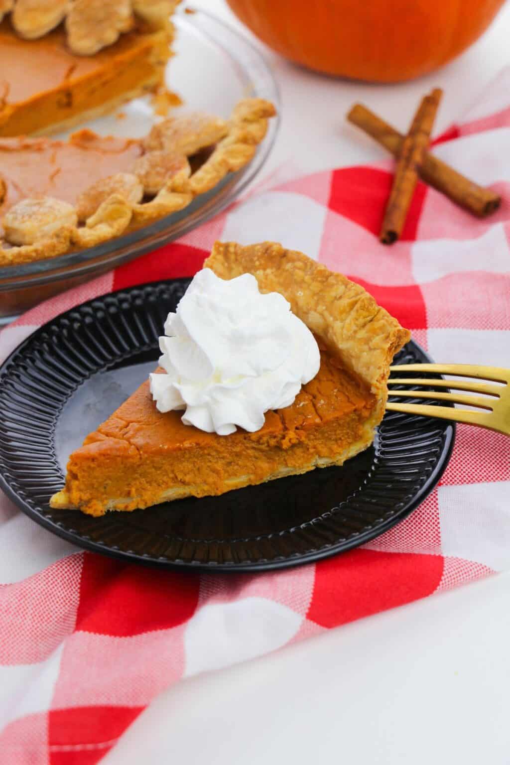 A slice of Old Fashioned Pumpkin Pie with whipped cream on a black plate, set on a red and white checkered cloth, with a gold fork and cinnamon sticks nearby.
