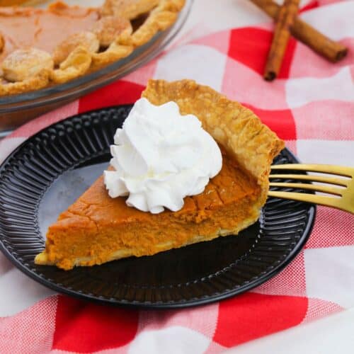 A slice of Old Fashioned Pumpkin Pie with whipped cream on a black plate, set on a red and white checkered cloth, with a gold fork and cinnamon sticks nearby.