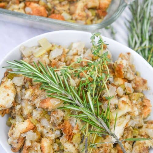 A bowl of Old Fashioned Stuffing with chopped vegetables, topped with fresh rosemary and thyme sprigs, sits next to a glass baking dish filled with stuffing.