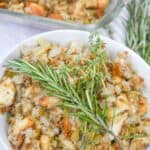 A bowl of Old Fashioned Stuffing topped with a sprig of fresh rosemary and thyme, with a glass baking dish of stuffing in the background.
