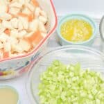 Bowls containing cubed bread, chopped celery, melted butter, and a small cup of liquid are arranged on a white surface.