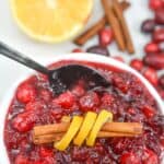 A bowl of Orange Cranberry Sauce garnished with a cinnamon stick and lemon peel, with a spoon inside; lemon half, cinnamon sticks, and fresh cranberries in the background.