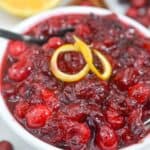 A bowl of relish with whole cranberries, garnished with lemon peel, sits invitingly with a spoon inside. Slices of orange and cinnamon sticks add a festive touch in the background.