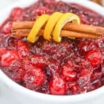 A bowl of Orange Cranberry Sauce garnished with a cinnamon stick and lemon peel, with a lemon half and cinnamon sticks in the background.