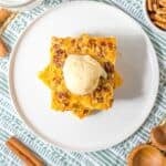 A square slice of Pumpkin Crunch Cake topped with ice cream sits on a white plate, surrounded by pecans, cinnamon sticks, and a glass of milk for the perfect autumn treat.