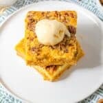 Two stacked Pumpkin Crunch Cake pecan dessert bars on a white plate, topped with a scoop of vanilla ice cream; cinnamon sticks and a cup are nearby.