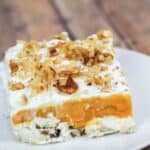 A square slice with a creamy white base, orange middle layer, whipped topping, and chopped walnuts, served on a white plate.