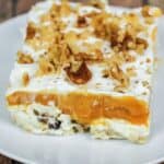 A square slice of Lush Cake featuring a creamy white base, bright orange layer, whipped topping, and chopped nuts is served on a white plate.