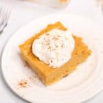 A square piece of Pumpkin Pie Dessert Bar topped with whipped cream and a sprinkle of cinnamon sits on a white plate.